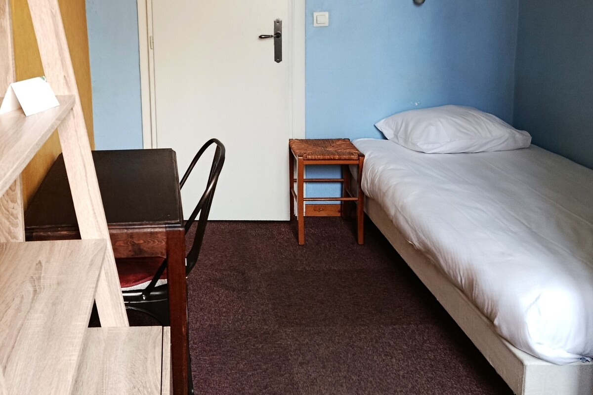 Una habitación con una cama, un escritorio, una silla, una puerta con un aviso y una lámpara fijada a la pared.
