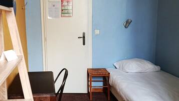 A room with a bed, a desk, a chair, a door with a notice, and a wall-mounted light fixture.