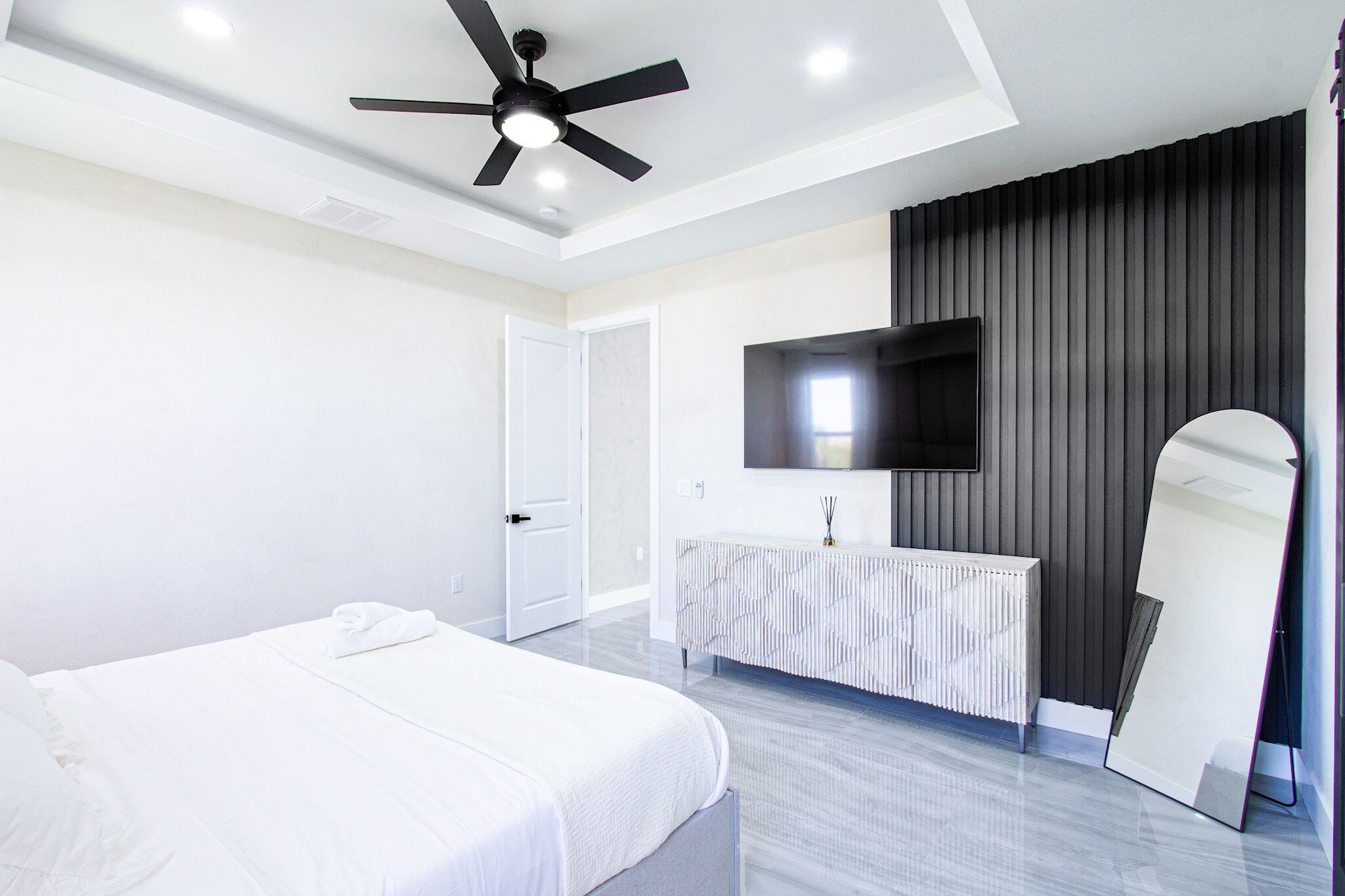 A modern hotel room with a bed, a ceiling fan, a flat-screen TV, a mirror, and a white console table.