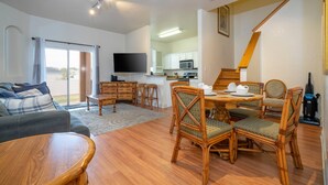 Amplia sala de estar con sofá, mesa de comedor y sillas, una cocina al fondo y una escalera.