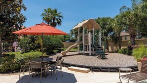 Un patio al aire libre con una mesa y sillas, un área de juegos con un tobogán y palmeras al fondo.
