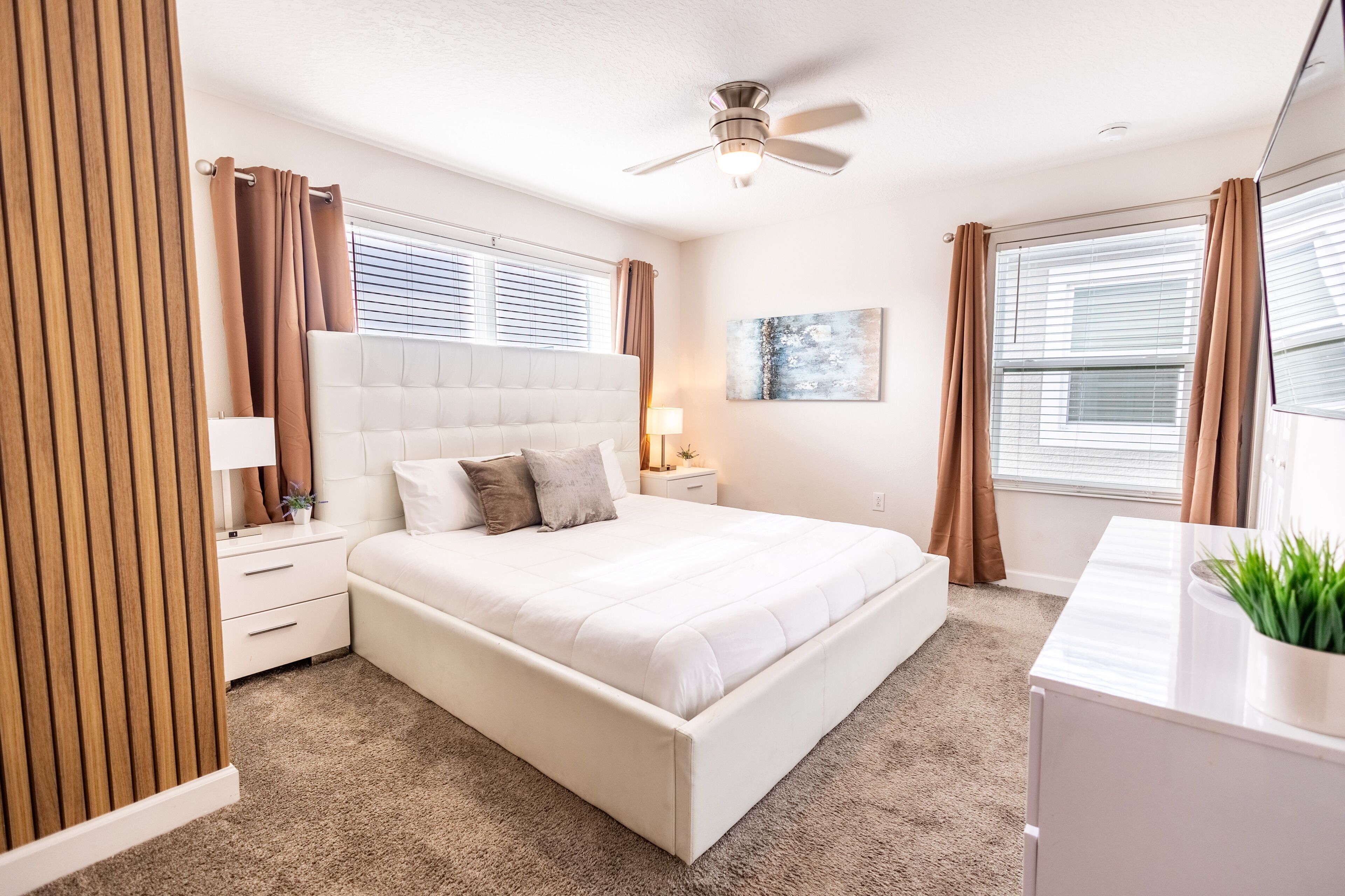 Une chambre à coucher avec un grand lit, un ventilateur de plafond, une table de chevet, une lampe et une fenêtre avec des rideaux.