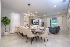 A modern dining area with a large table set for six, surrounded by six chairs. There is a television mounted on the wall, a potted plant, and a sofa in the background.