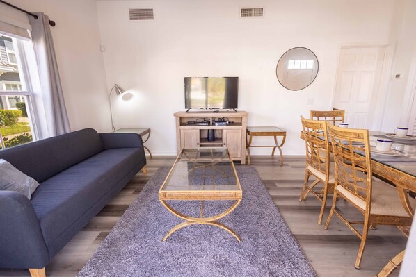 A modern living room with a sofa, a glass-top coffee table, a TV, and a dining area with wooden chairs and a table.