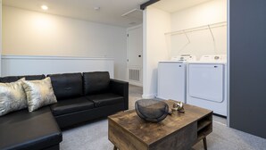 A modern living room with a black leather sofa, a wooden coffee table, and a washing machine in the background.