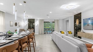 Un espace repas moderne avec une longue table, des chaises et des luminaires suspendus. On y trouve également un coin salon avec un canapé et des coussins.