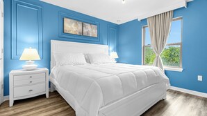 Une chambre avec un lit blanc, deux tables de chevet avec des lampes, une fenêtre avec des rideaux et un mur bleu orné d’un tableau au-dessus du lit.