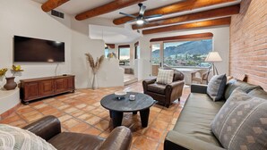 A spacious living area with a large flat-screen TV, leather sofas, a coffee table, and a view of a mountainous landscape through large windows. - Cimarron: Spectacular city and mountain views, pool, hot tub, and more! (Catalina Foothills)