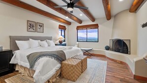 A bedroom with a large bed, a ceiling fan, wooden beams, a fireplace, and a view of the landscape. - Cimarron: Spectacular city and mountain views, pool, hot tub, and more! (Catalina Foothills)