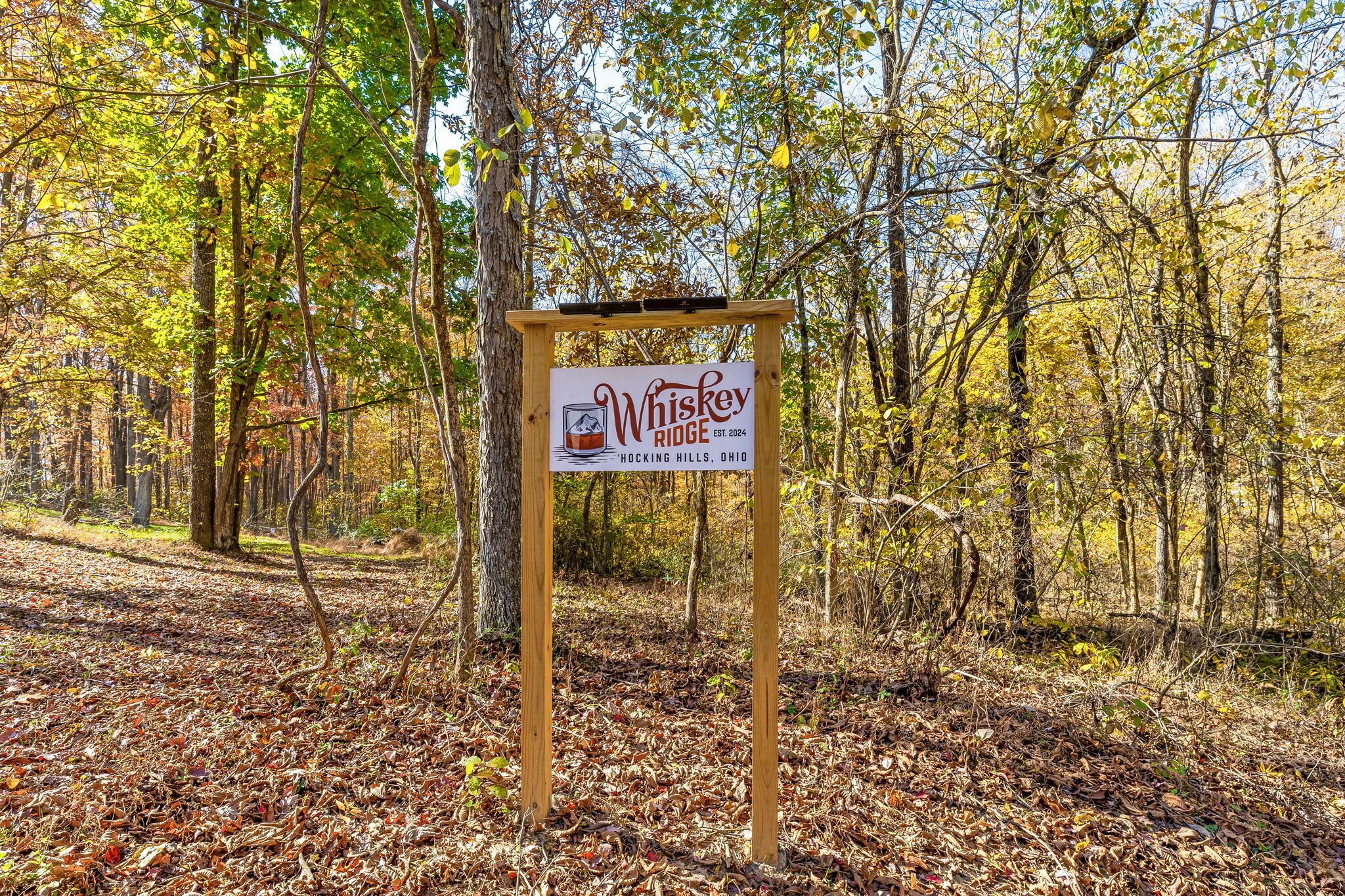A wooden sign in a forested area with a whiskey logo and text indicating it is Whiskey Ridge, established in 2004, located in Hocking Hills, Ohio.