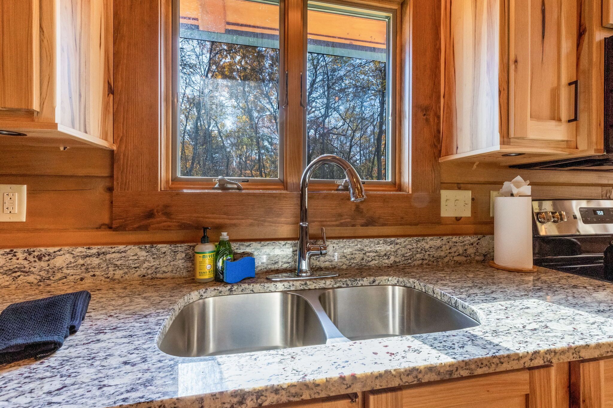A kitchen with wooden cabinets, a granite countertop, a stainless steel sink, and a window with a view of trees.