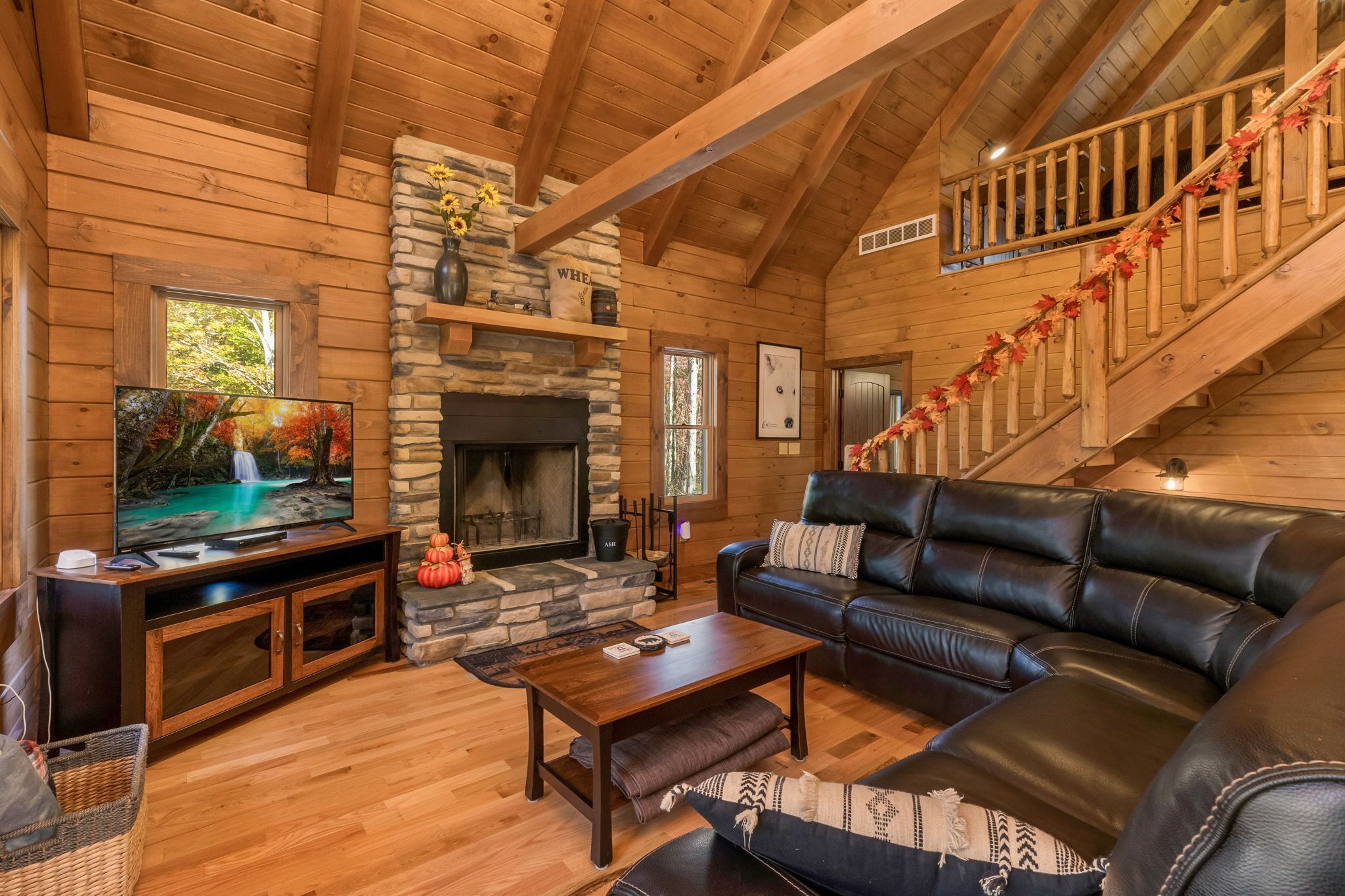 A cozy log cabin interior with a stone fireplace, a leather sofa, a television, and a staircase with wooden railings.