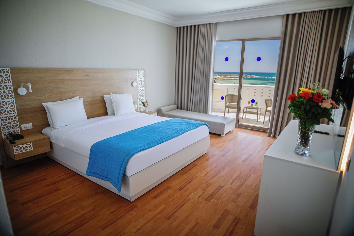 Ein Hotelzimmer mit einem großen Bett, einer Couch, einem Schreibtisch, einem Fernseher und einem Balkon mit Meerblick.