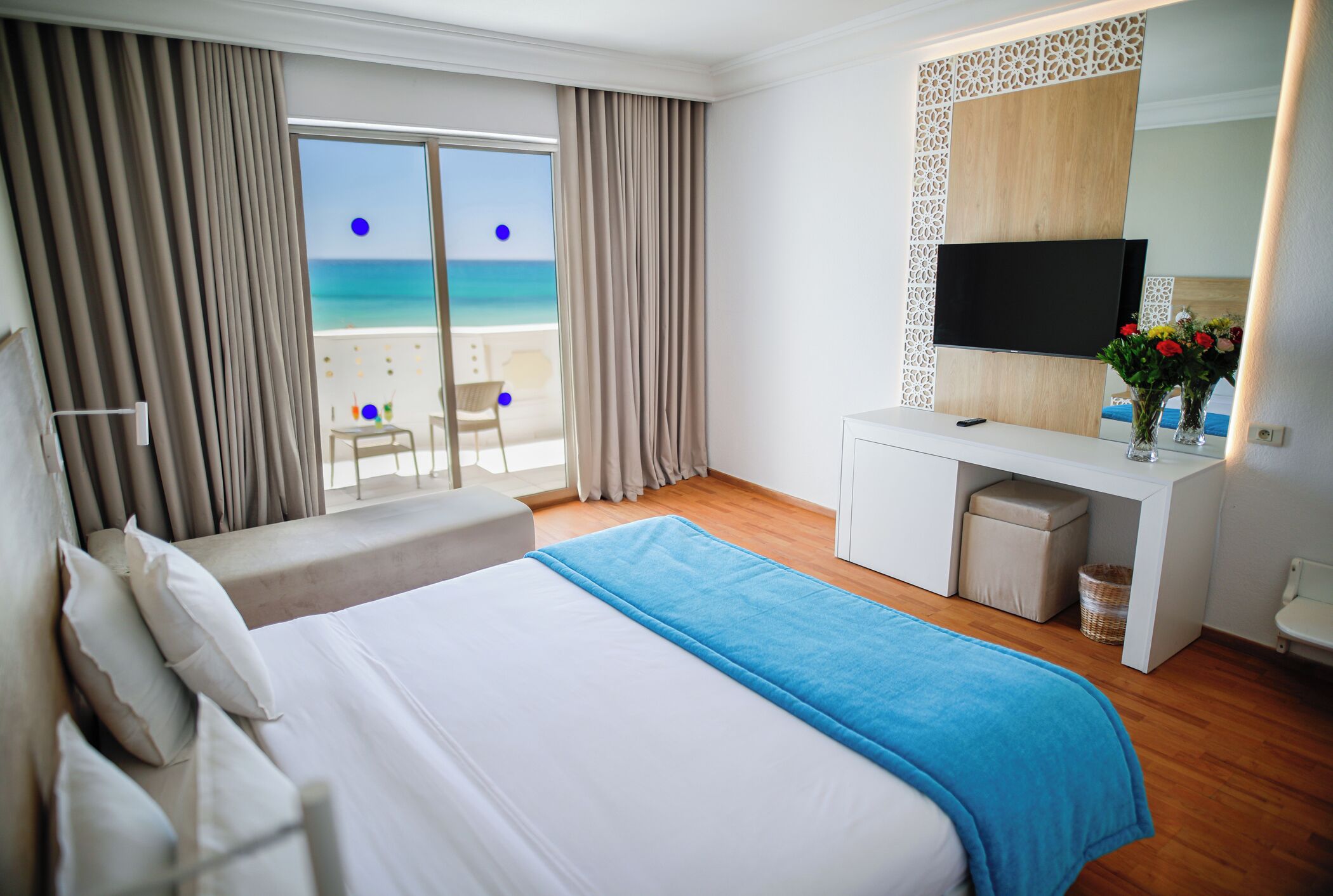 A hotel room with a bed, a desk with a TV, a view of the beach, and a balcony.
