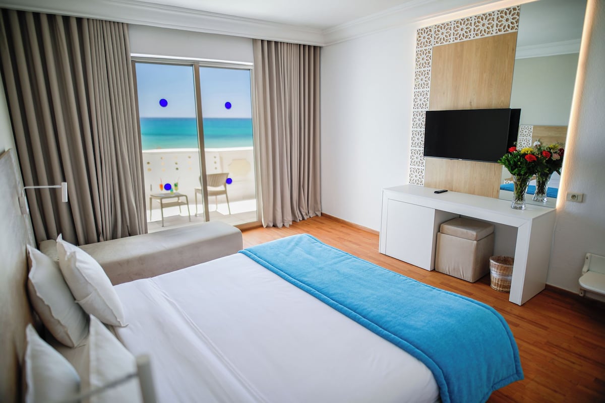 A hotel room with a bed, a desk with a TV, a view of the beach, and a balcony.