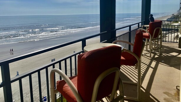 A balcony with a view of the beach, featuring chairs and a table. - Sea Viewers Dream Oceanfront 5th floor ~ Kitchen Living room Bedroom  Pools Open (Daytona Beach)