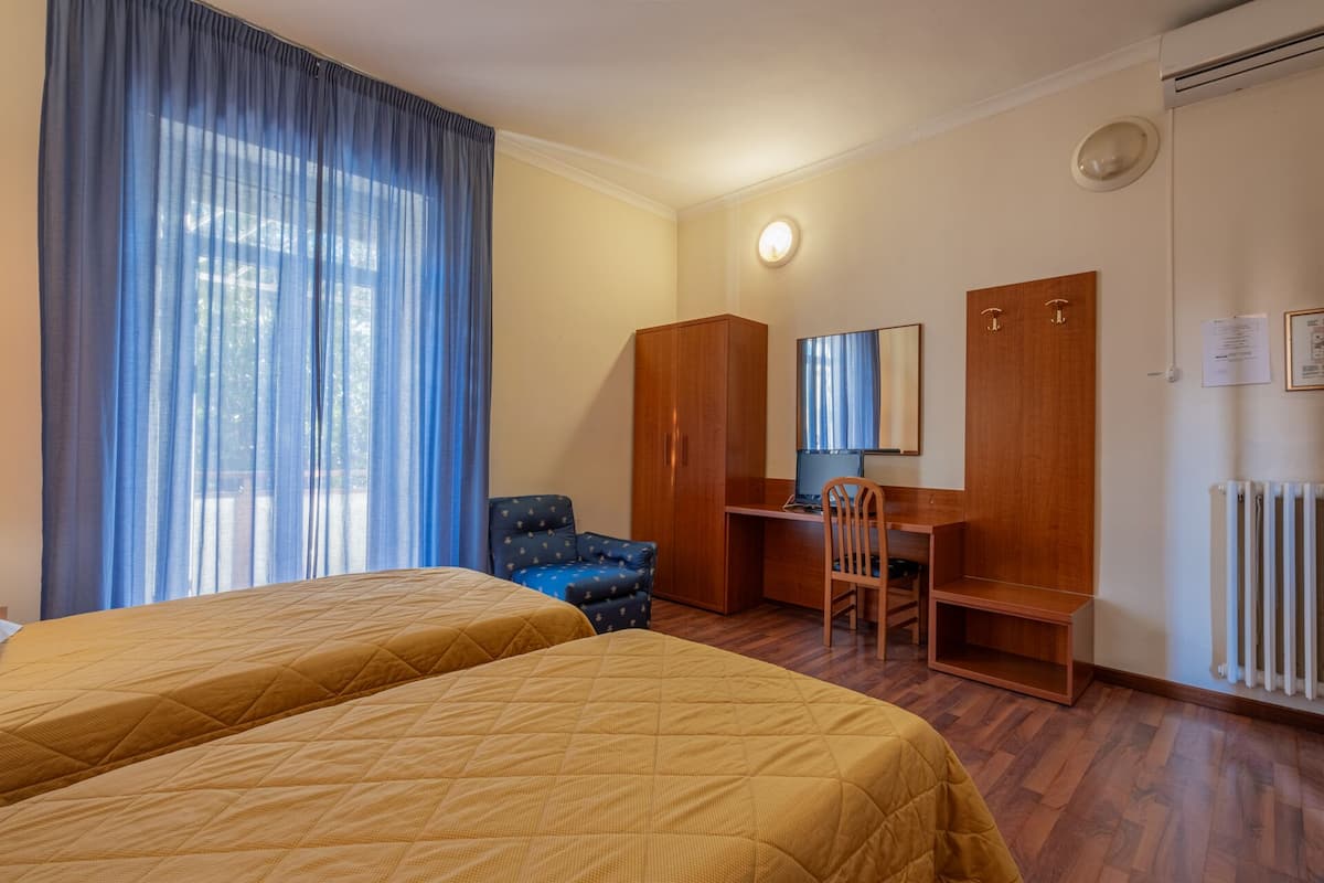 Habitación de hotel con dos camas, un escritorio con computadora, una silla, un armario y un radiador.