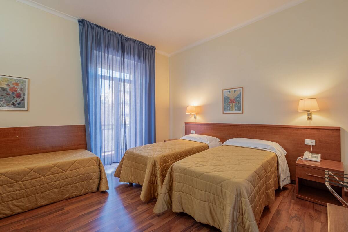 Habitación de hotel con dos camas individuales, cabeceros de madera, un ventanal grande con cortinas azules y dos lámparas de pared.