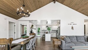 A spacious living area with a high ceiling, a dining table with chairs, a grey sofa, and a surfboard on a side table. - The Cottage at Grand Marais (Grand Marais)