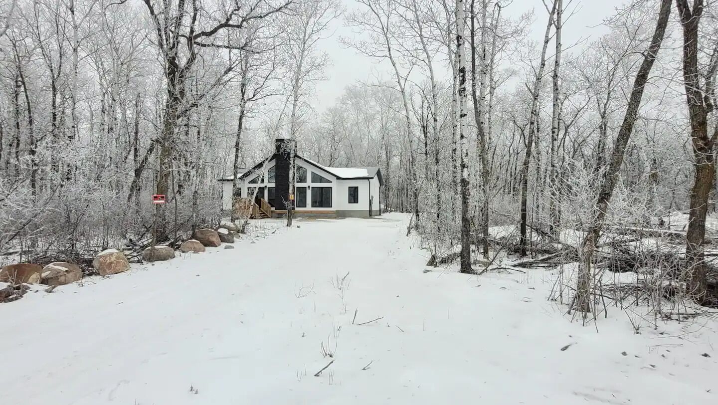 Un sentier enneigé mène à une cabane blanche au toit noir, entourée d’arbres et de rochers.