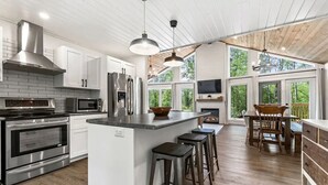 A modern kitchen with stainless steel appliances, a central island with stools, and a dining area with a view of the outdoors. - The Cottage at Grand Marais (Grand Marais)