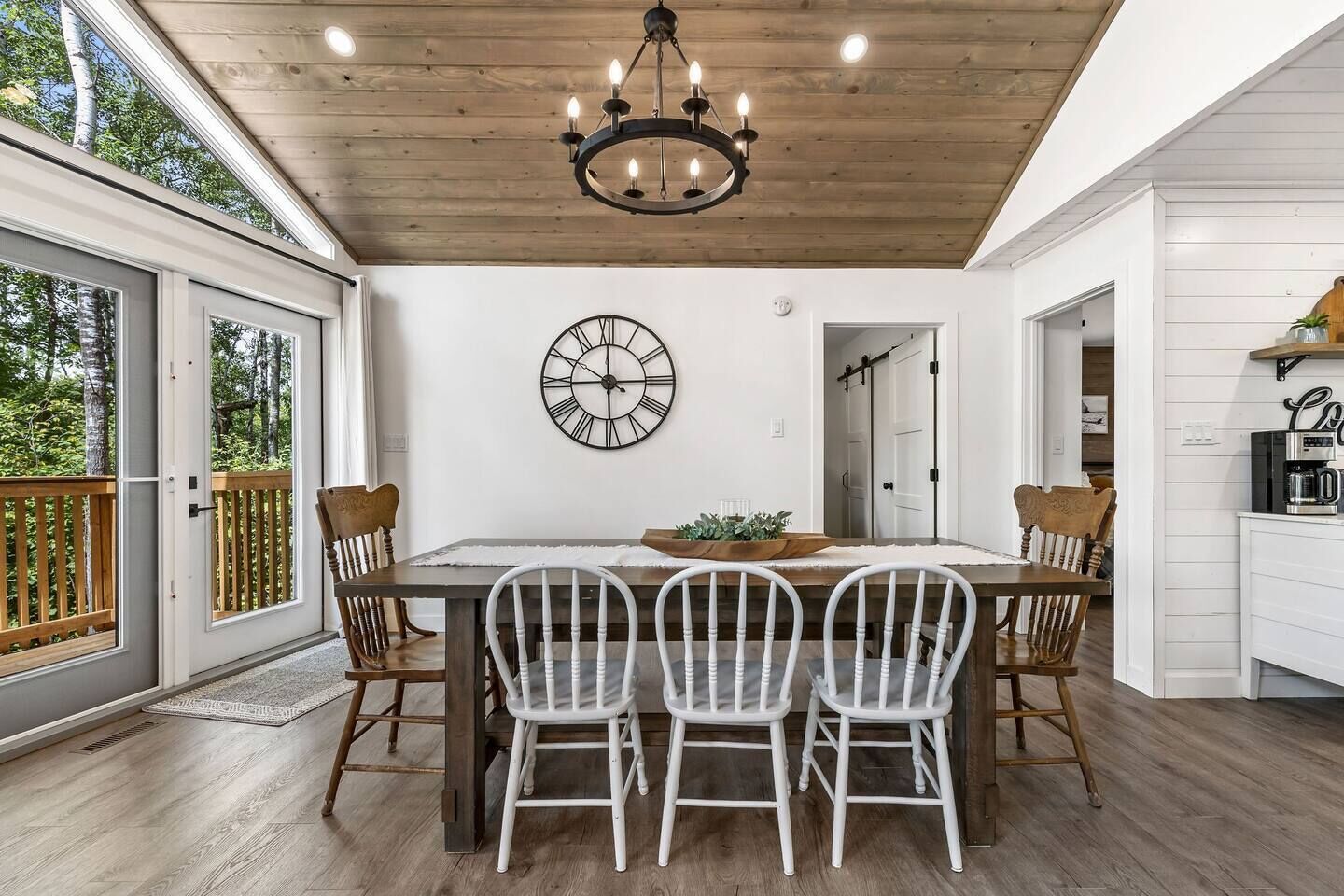 A dining area with a wooden table, white chairs, a large wall clock, and a chandelier.