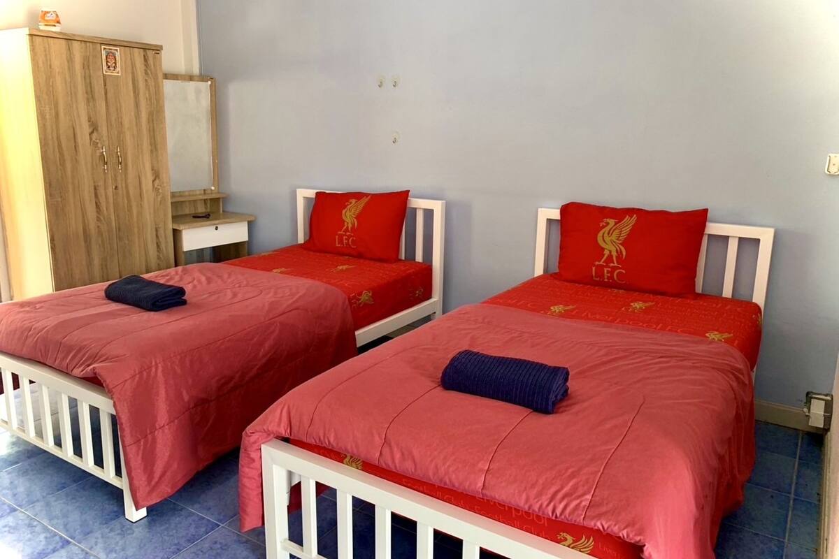 Duas camas de solteiro com roupa de cama e almofadas vermelhas, um guarda-roupa de madeira e uma mesa pequena.