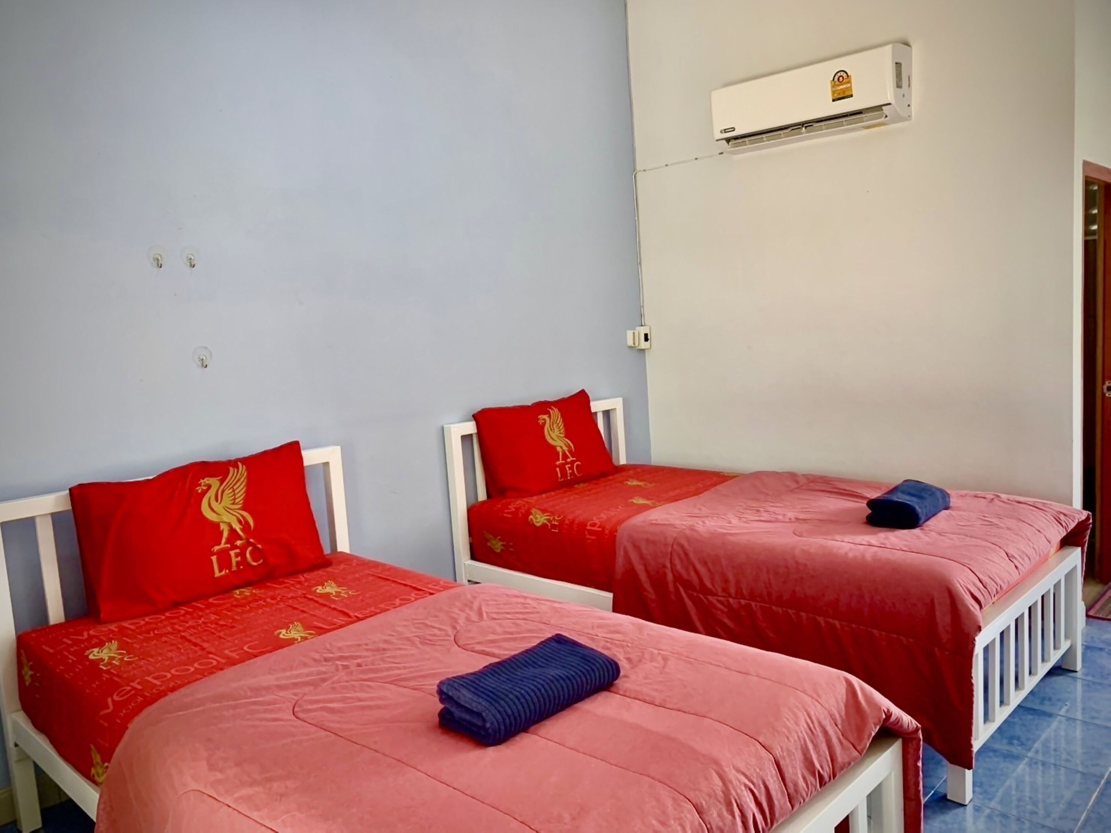 Una stanza con due letti singoli, ognuno con una coperta rossa e un cuscino con lo stemma di un leone dorato. Su un letto, un asciugamano blu.