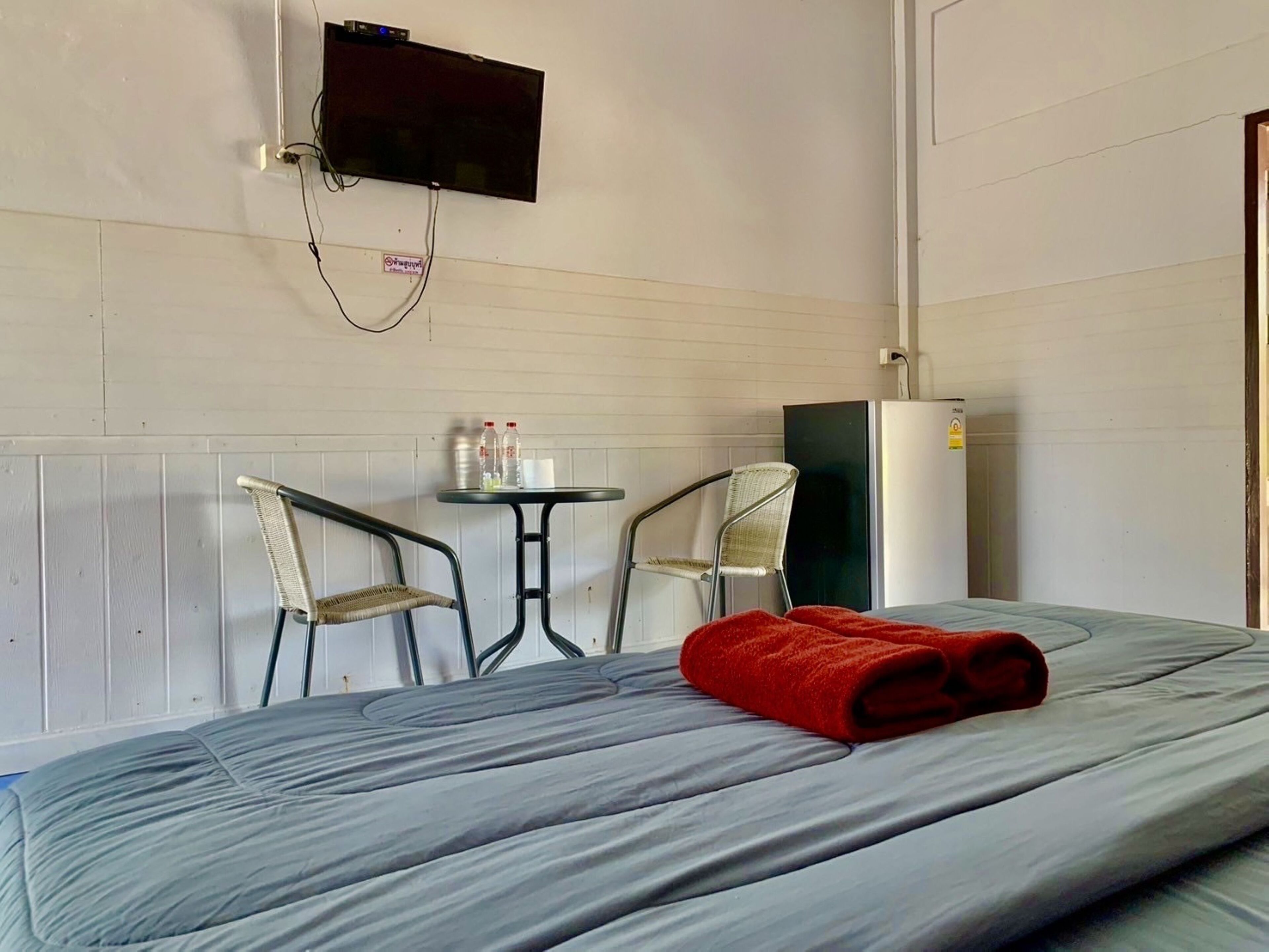 A single bed with a grey mattress and a red towel, two metal chairs, a small round table with bottles and a cup, a black TV mounted on the wall, and a white refrigerator.