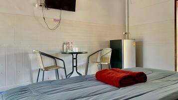 A single bed with a grey mattress and a red towel, two metal chairs, a small round table with bottles and a cup, a black TV mounted on the wall, and a white refrigerator.