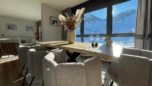 A dining area with a wooden table, grey upholstered chairs, a vase with dried plants, and a view of snow-capped mountains through large windows. - Chez Tipère à La Mongie (Bagnères-de-Bigorre)