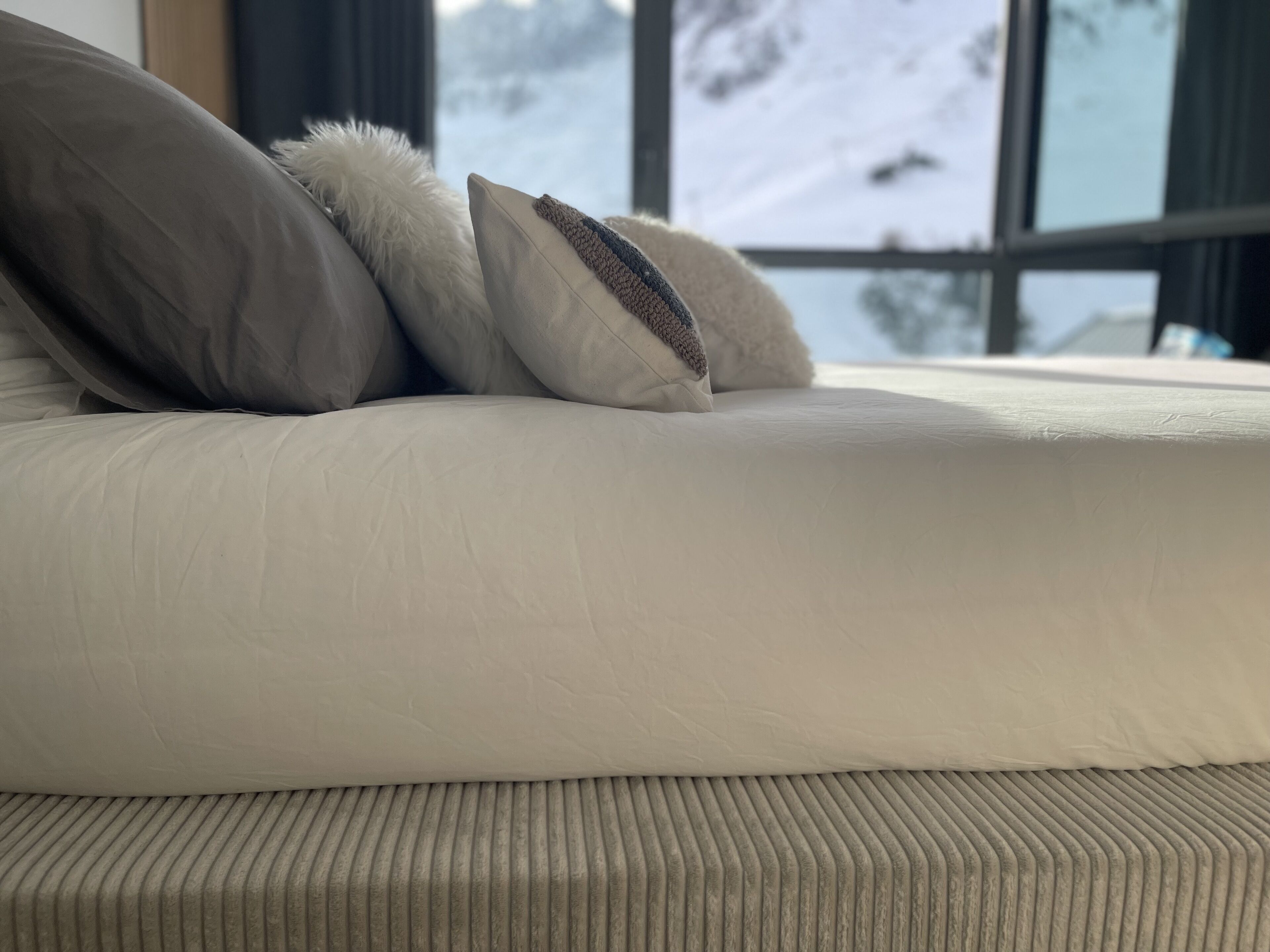 Una cama con colchón blanco y almohadas grises, ubicada cerca de una ventana con vistas a un paisaje nevado.