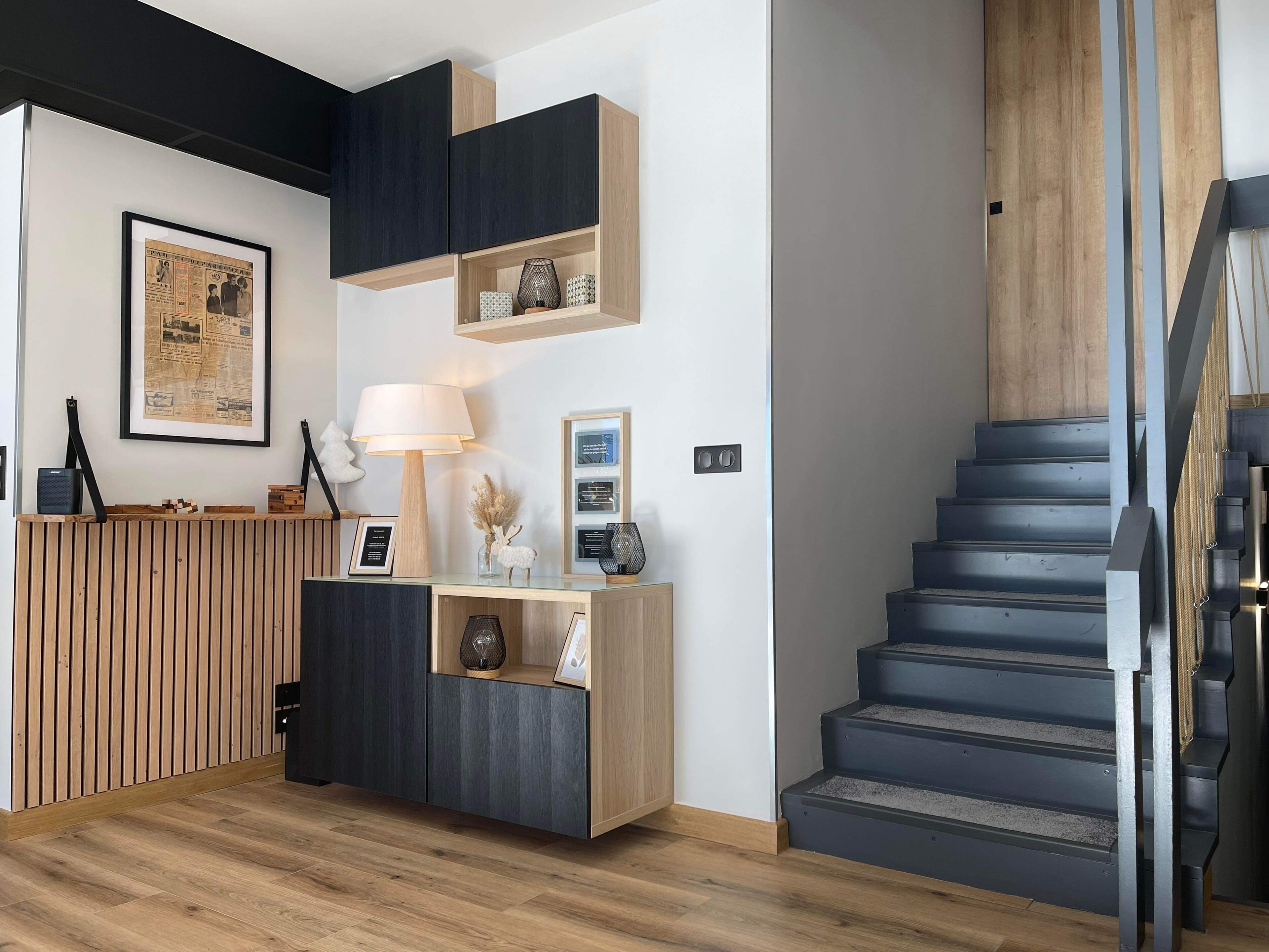 Un intérieur moderne avec un comptoir d’accueil en bois, un journal encadré, une lampe et un escalier.
