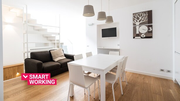 Une pièce moderne et bien éclairée, avec une table blanche, des chaises, un canapé noir et un escalier.