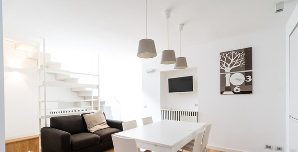 A modern, well-lit room with a white table, chairs, a black sofa, and a staircase.