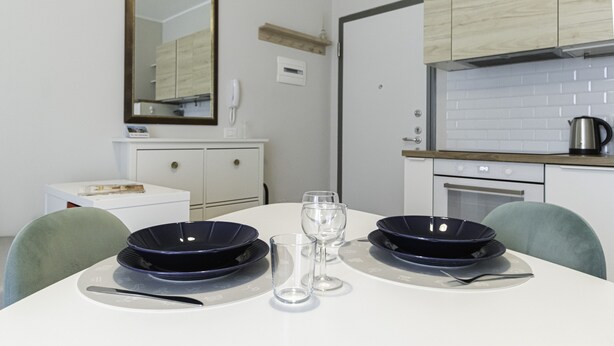A dining area with a white table set for two, two dark blue plates, two clear wine glasses, and a knife. - Italianway Easy - Tartini 23 (Milano)