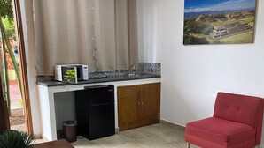 Une cuisine compacte équipée d’un four à micro-ondes, d’un réfrigérateur et d’un évier. On y trouve une chaise rouge et une table à manger agrémentée d’une plante en pot. Un tableau représentant un paysage est accroché au mur.