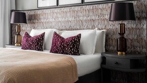 Un lit bien fait, avec des oreillers blancs et violets à motifs, une couverture beige et deux lampes de chevet posées sur une table de nuit sombre.