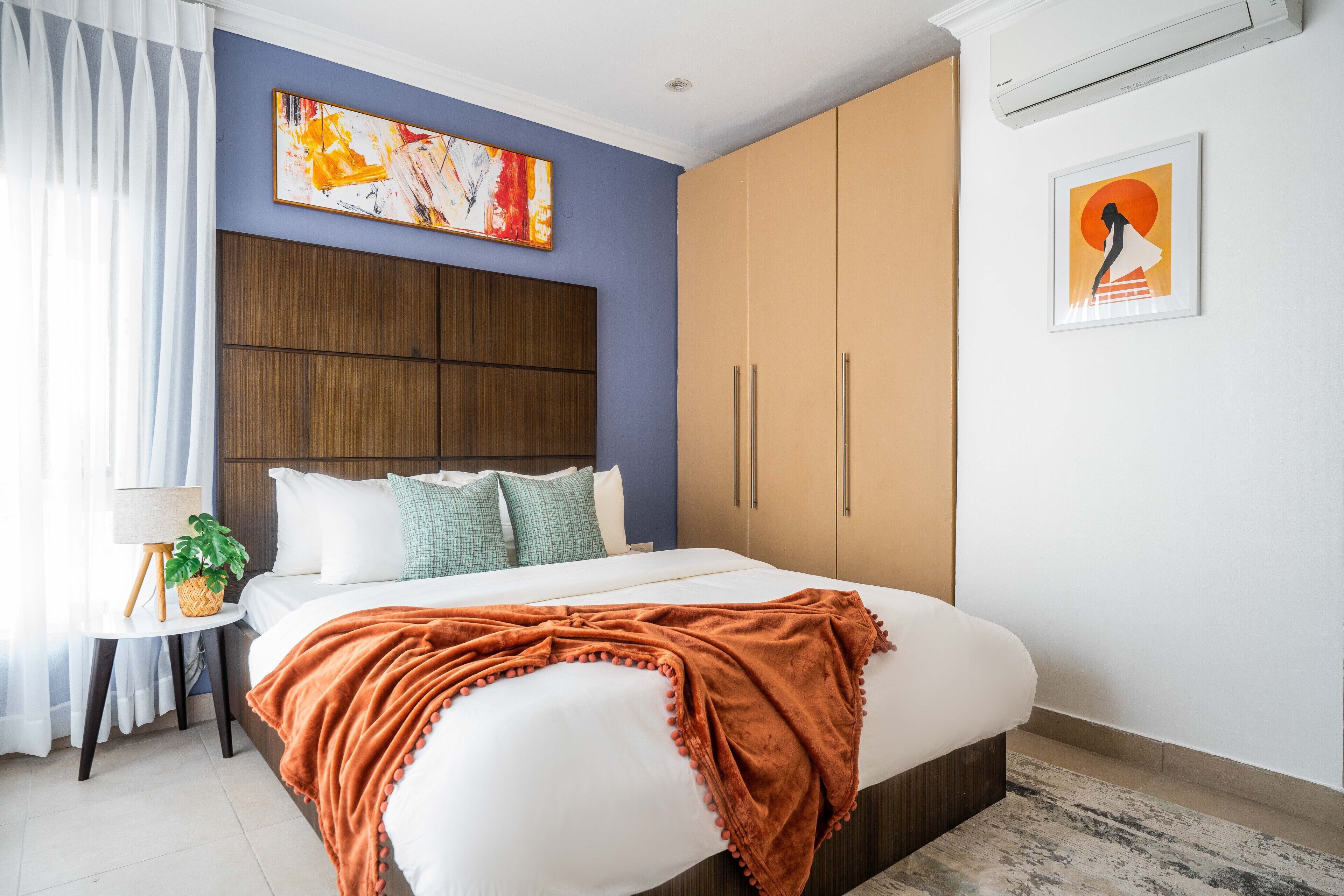A bedroom with a bed, a nightstand, a lamp, a potted plant, and artwork on the walls.