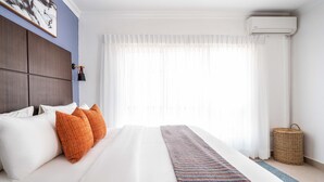 Een net opgemaakt bed met witte en oranje kussens, een gestreept dekbed en een hoofdbord tegen een blauwe muur.