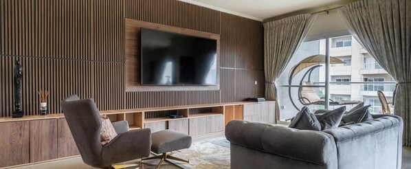 A modern living room with a large flat-screen TV mounted on a wooden wall, a grey sofa, and a chair with a cushion.