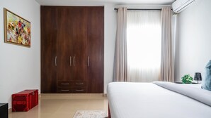 Une chambre à coucher avec un lit, une armoire, une valise rouge, une plante et un tableau accroché au mur.