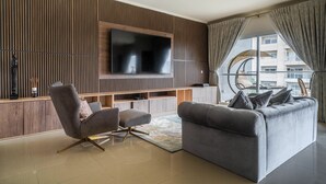 Un salon moderne doté d’un grand téléviseur à écran plat fixé sur un mur recouvert de panneaux en bois, d’un canapé gris et d’un fauteuil gris assorti, accompagné d’un pouf.