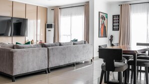 Un salon moderne avec un canapé d’angle gris, un téléviseur à écran plat, une table à manger avec des chaises et une grande fenêtre avec des rideaux.