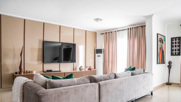 Un salon moderne avec un grand canapé d’angle, un téléviseur à écran plat fixé au mur et une grande fenêtre agrémentée de rideaux.