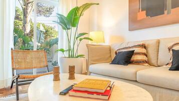 Een woonkamer met een bank, een bijzettafeltje met boeken, een lamp, een stoel en een grote plant.