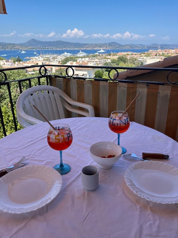 A table set for two with wine glasses, plates, and a bowl on a balcony overlooking a coastal cityscape.