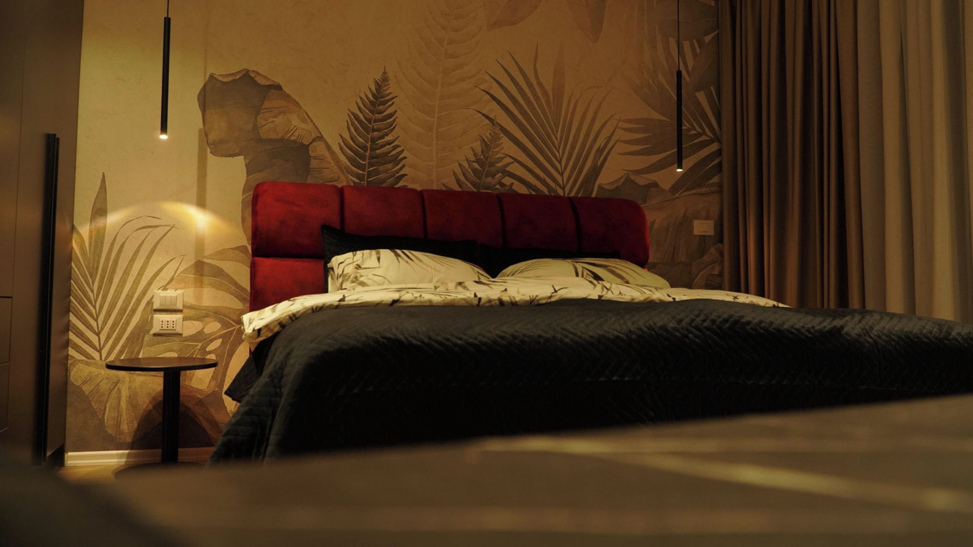 A bedroom with a red upholstered headboard, a bed with patterned bedding, a nightstand with a lamp, and a wall with a leaf pattern.