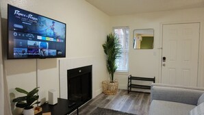 Een woonkamer met een flatscreen televisie waarop 'Prince of Persia: The Lost Crown' draait, een open haard, een grijze bank, een klein tafeltje met een plant en een deur op de achtergrond.
