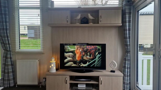 A living room with a TV on a wooden stand, a small table with a lamp, and a window with blinds. - Inviting 2-bed Caravan on Combe Haven Holiday Park (St Leonards-On-Sea)